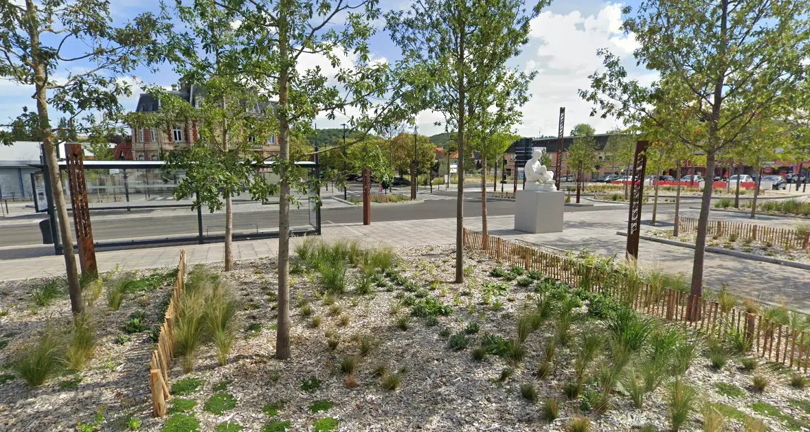 Vue d'une place aménagée avec des jeunes arbres, des parterres de plantes et des clôtures en bois, bordée par un arrêt de bus moderne. Au centre, une statue blanche repose sur un socle, avec des bâtiments et une rue en arrière-plan