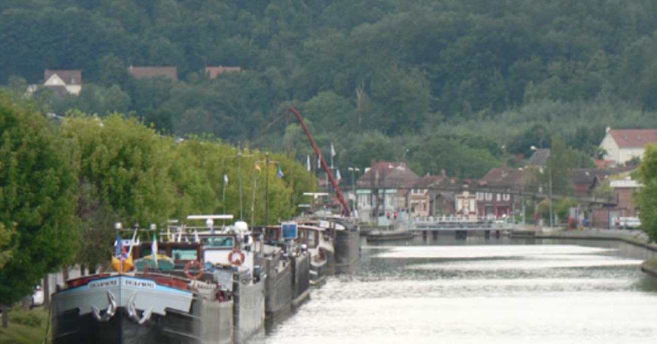 Bateaux amarrés le long d'un canal bordé d'arbres et de maisons, avec une forêt verdoyante en arrière-plan dans l'Oise (60)