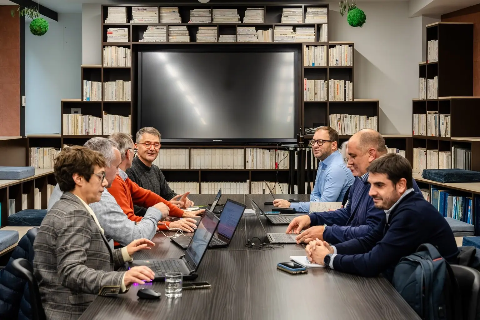 Groupe de personnes en réunion autour d'une grande table, dans une bibliothèque moderne équipée d'un écran