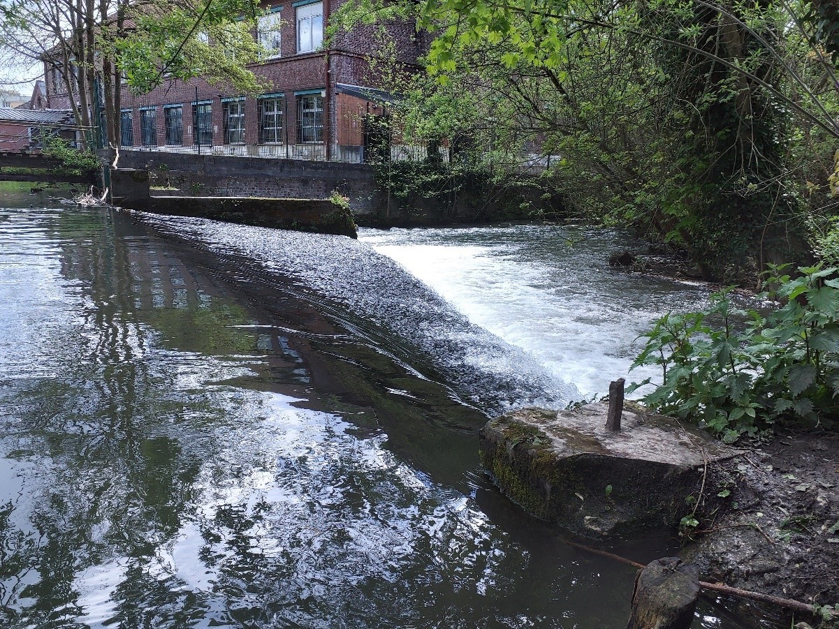 Vue d’un ancien bâtiment industriel en briques rouges bordant une rivière, avec un petit barrage en déversoir d’eau créant un léger courant et des remous. La végétation dense entoure la rive, avec des arbres et arbustes proches de l’eau