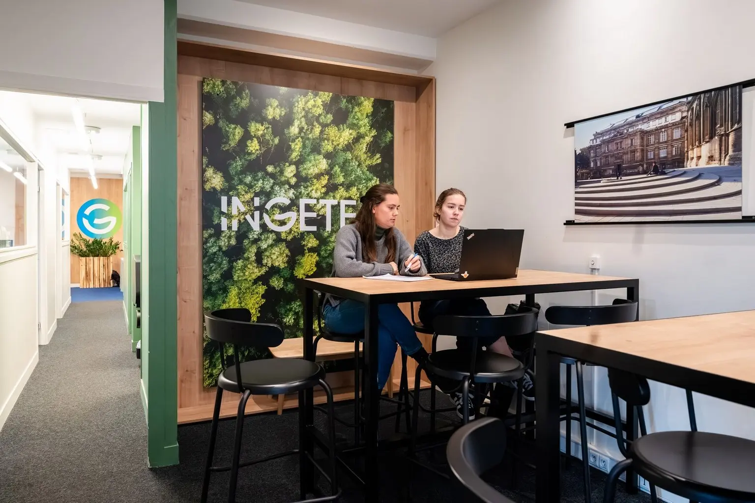 Deux jeunes femmes travaillant sur un ordinateur portable dans un espace moderne décoré d'une affiche végétale portant le logo 'INGETEC'