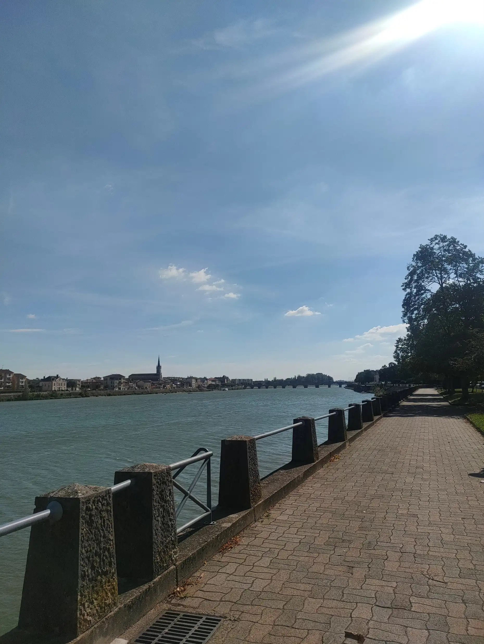 Vue d'une promenade pavée longeant un fleuve sous un ciel ensoleillé. Une rangée d'arbres à droite, des bâtiments au loin, et une église visible sur l'horizon