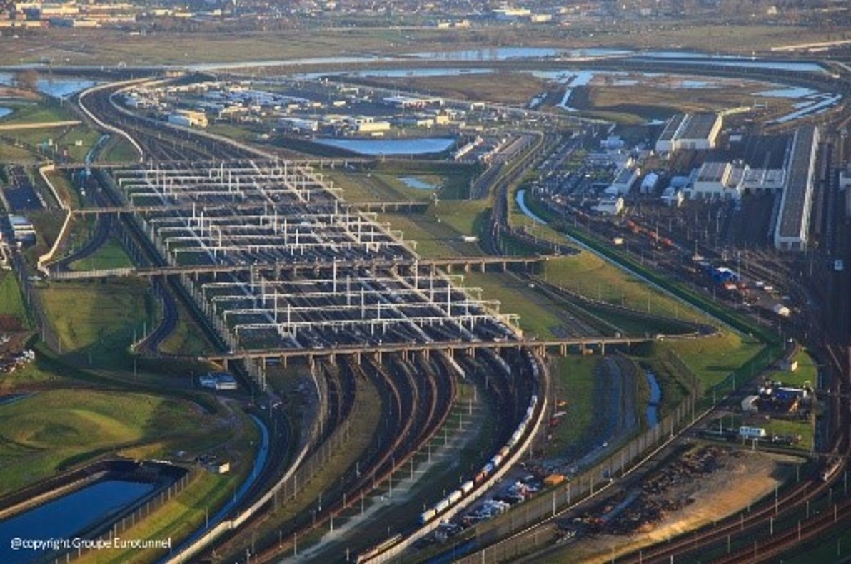 Vue aérienne du terminal Eurotunnel avec un réseau dense de rails et de ponts, de nombreuses voies ferrées alignées pour accueillir les trains sous une lumière de fin de journée