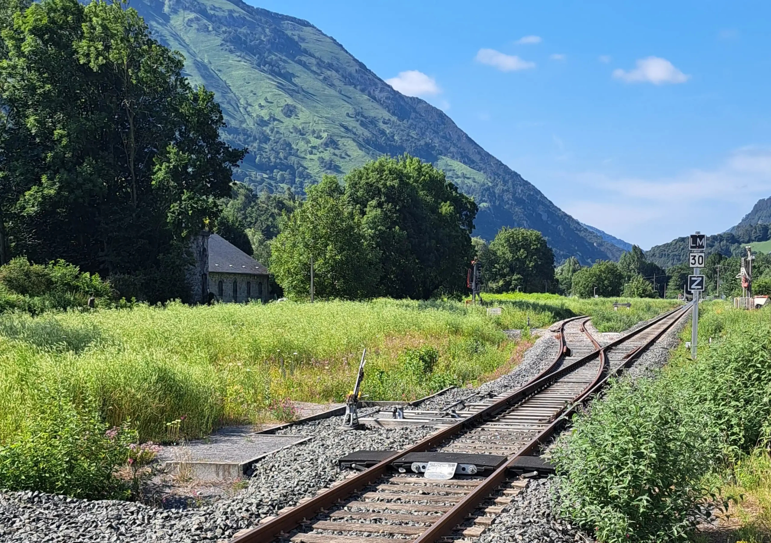 Voie ferrée en pleine nature, entourée de champs verdoyants et de montagnes en arrière-plan sous un ciel bleu. Une petite bâtisse en pierre est visible à gauche, partiellement cachée par des arbres