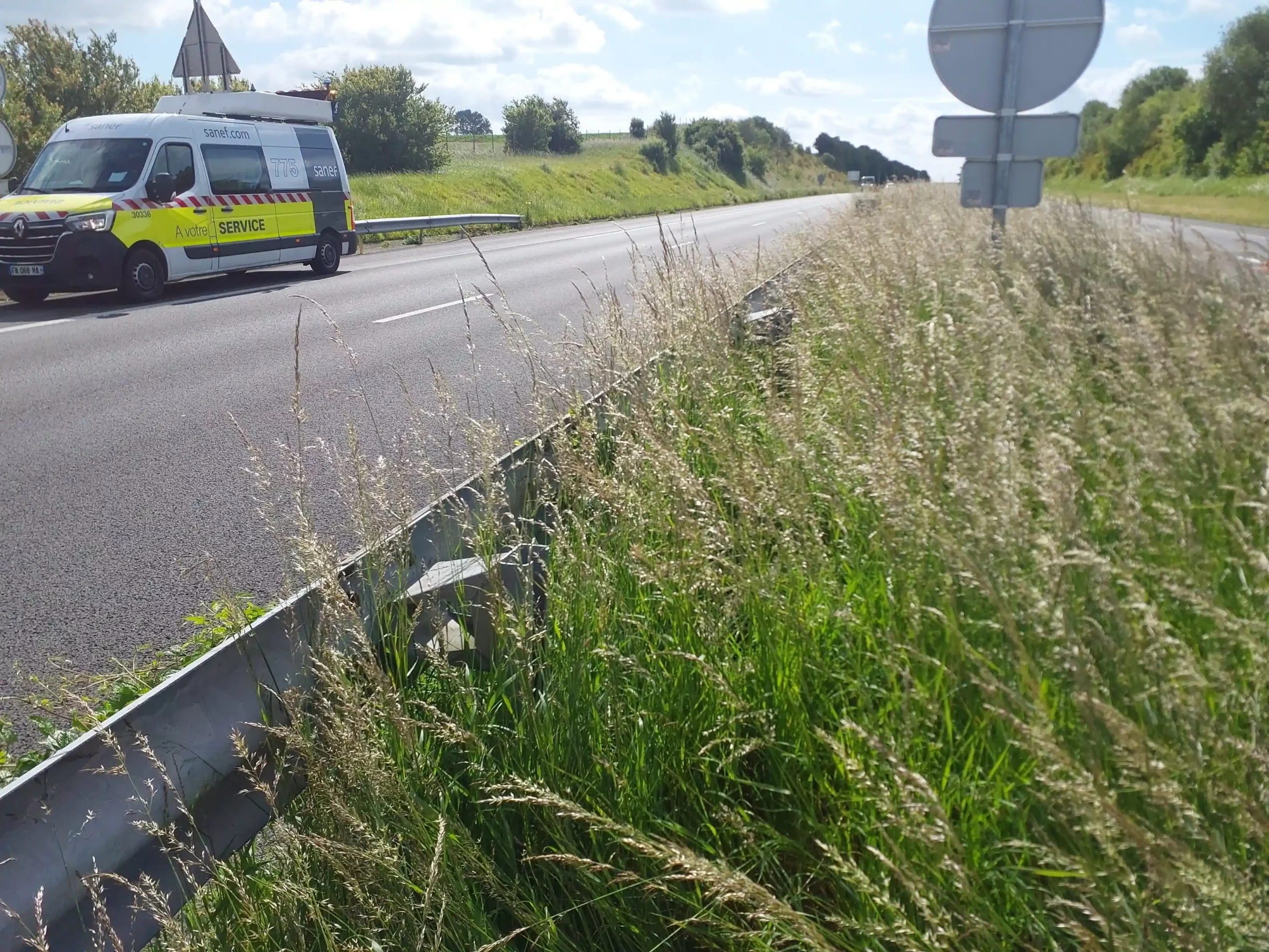 Photo illustrant une inspection d'ouvrage le long d'une route, avec un véhicule de service SANEF stationné et des herbes hautes au premier plan.