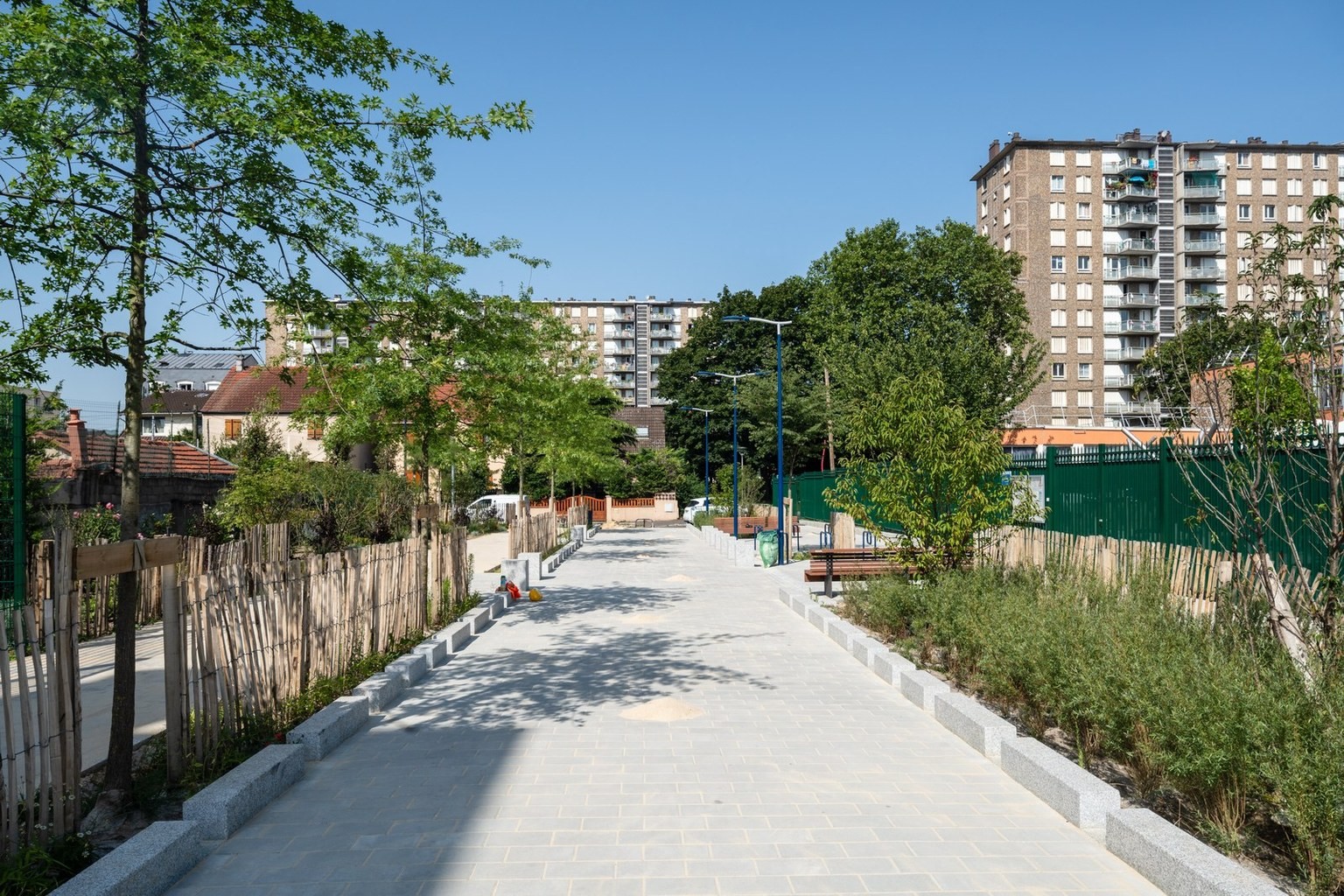 Vue d'un aménagement urbain à Drancy, avec une allée pavée bordée d'arbres, de bancs et d'espaces verts, entourée d'immeubles résidentiels.