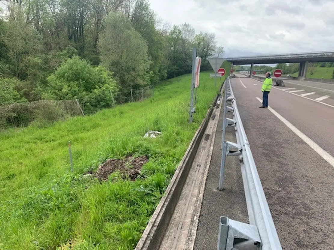 Photo montrant une voie d'autoroute avec un terre-plein en herbe à gauche, des panneaux de signalisation, et une personne portant une veste de sécurité.